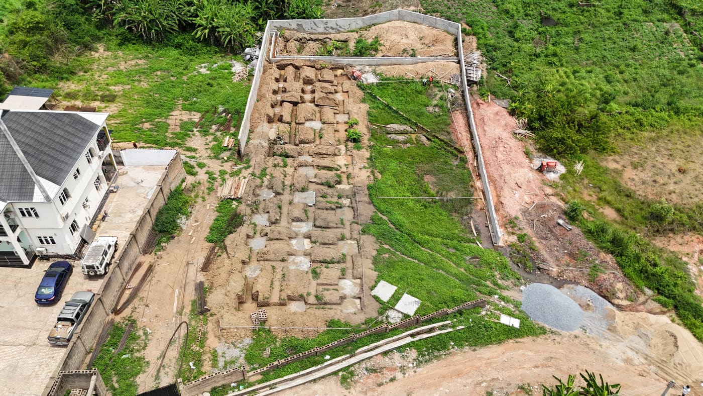 Ewang Estate under construction, 22 April 2025: the perimeter wall complete and the foundations set out — excavated trenches and cast pad footings across the cleared site, before any blockwork. Aerial view 1 of 2.