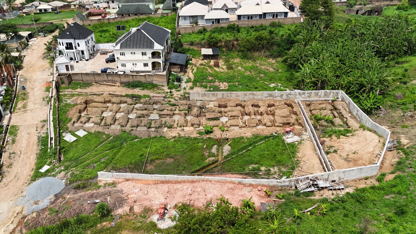Ewang Estate under construction, 22 April 2025: the perimeter wall complete and the foundations set out — excavated trenches and cast pad footings across the cleared site, before any blockwork. Aerial view 2 of 2.