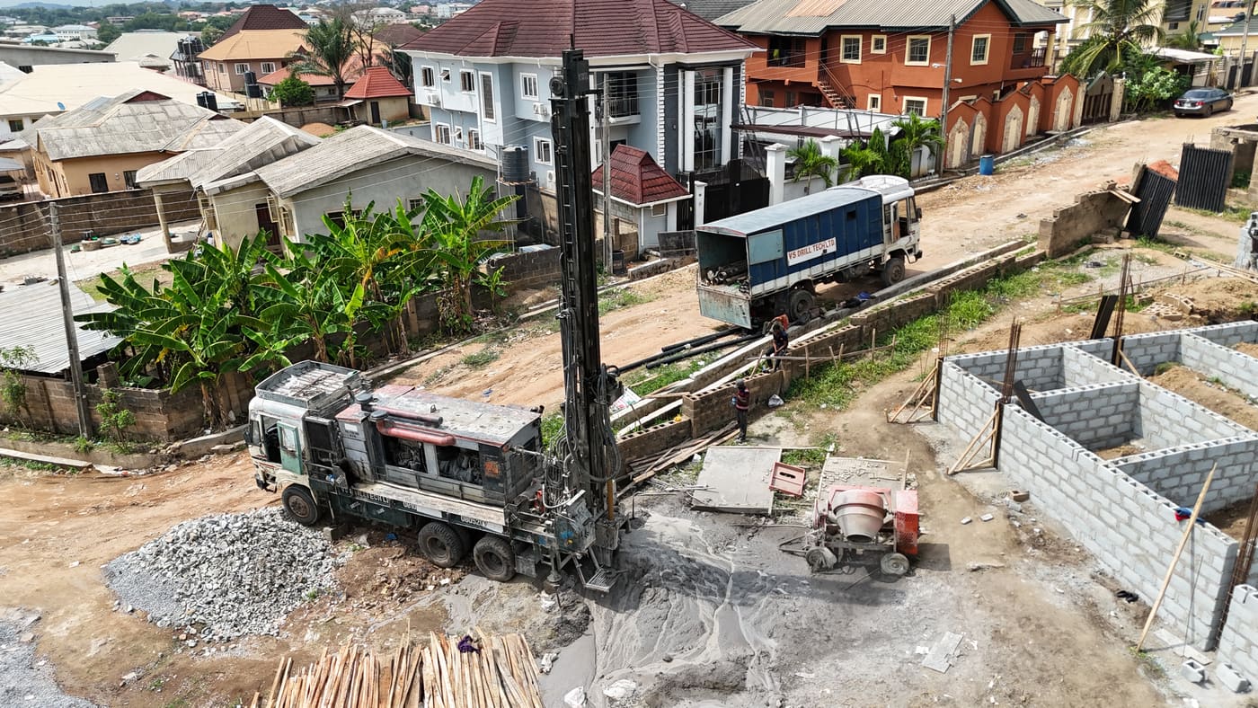 Ewang Estate under construction, 13 May 2025: a borehole rig drilling on site while the first foundation blockwork rises, with room divisions already readable at ground level. Aerial view 1 of 3.