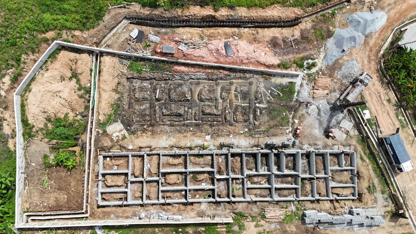 Ewang Estate under construction, 13 May 2025: a borehole rig drilling on site while the first foundation blockwork rises, with room divisions already readable at ground level. Aerial view 3 of 3.