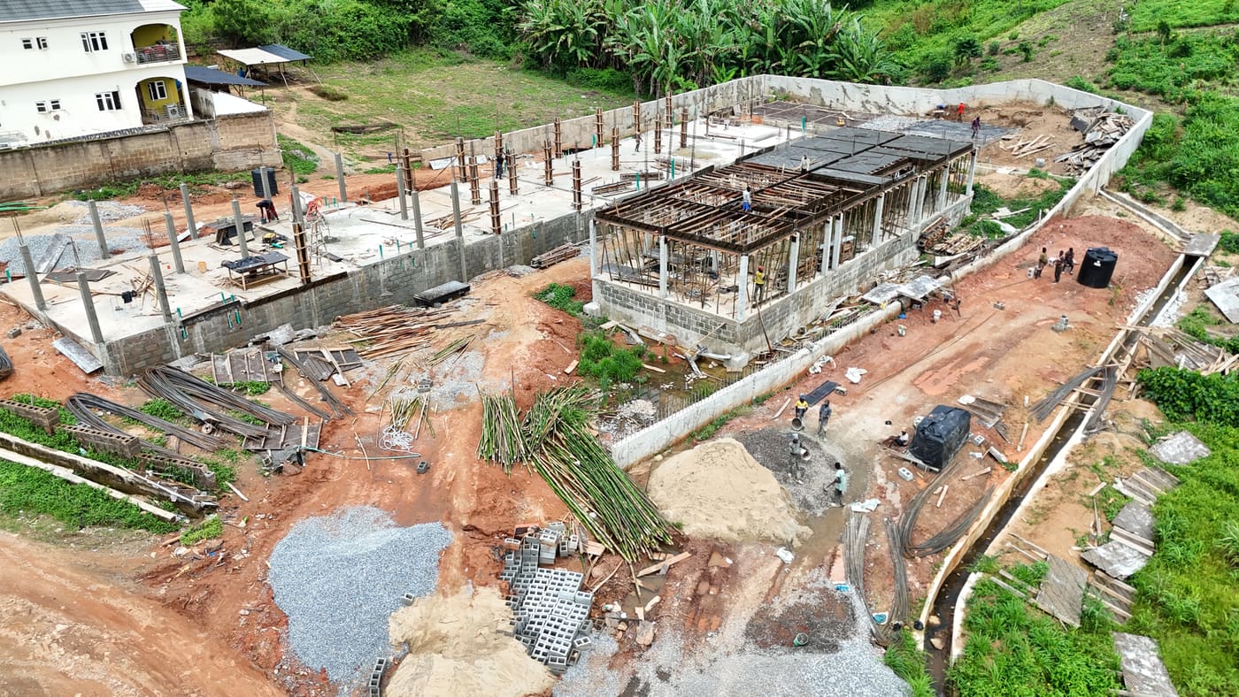 Ewang Estate under construction, 10 July 2025: the ground-floor slab cast and columns standing, with timber formwork and bamboo props carrying the first suspended slab as workers pour. Aerial view 1 of 4.