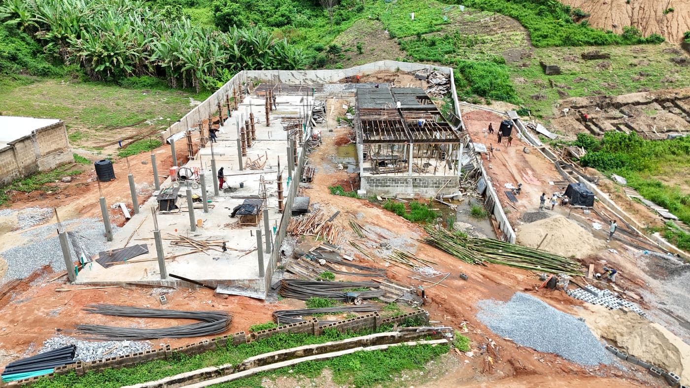 Ewang Estate under construction, 10 July 2025: the ground-floor slab cast and columns standing, with timber formwork and bamboo props carrying the first suspended slab as workers pour. Aerial view 2 of 4.
