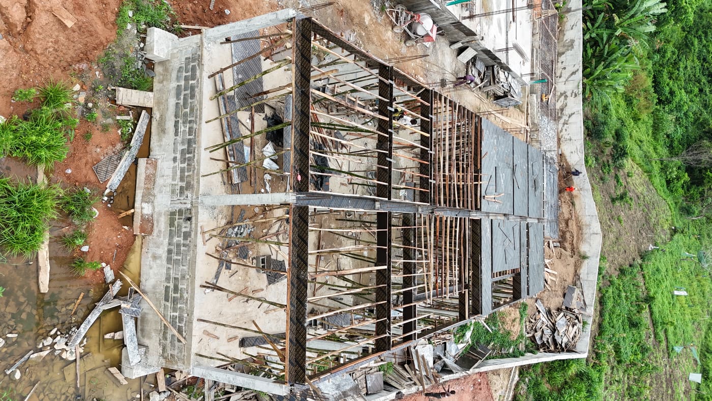 Ewang Estate under construction, 10 July 2025: the ground-floor slab cast and columns standing, with timber formwork and bamboo props carrying the first suspended slab as workers pour. Aerial view 3 of 4.