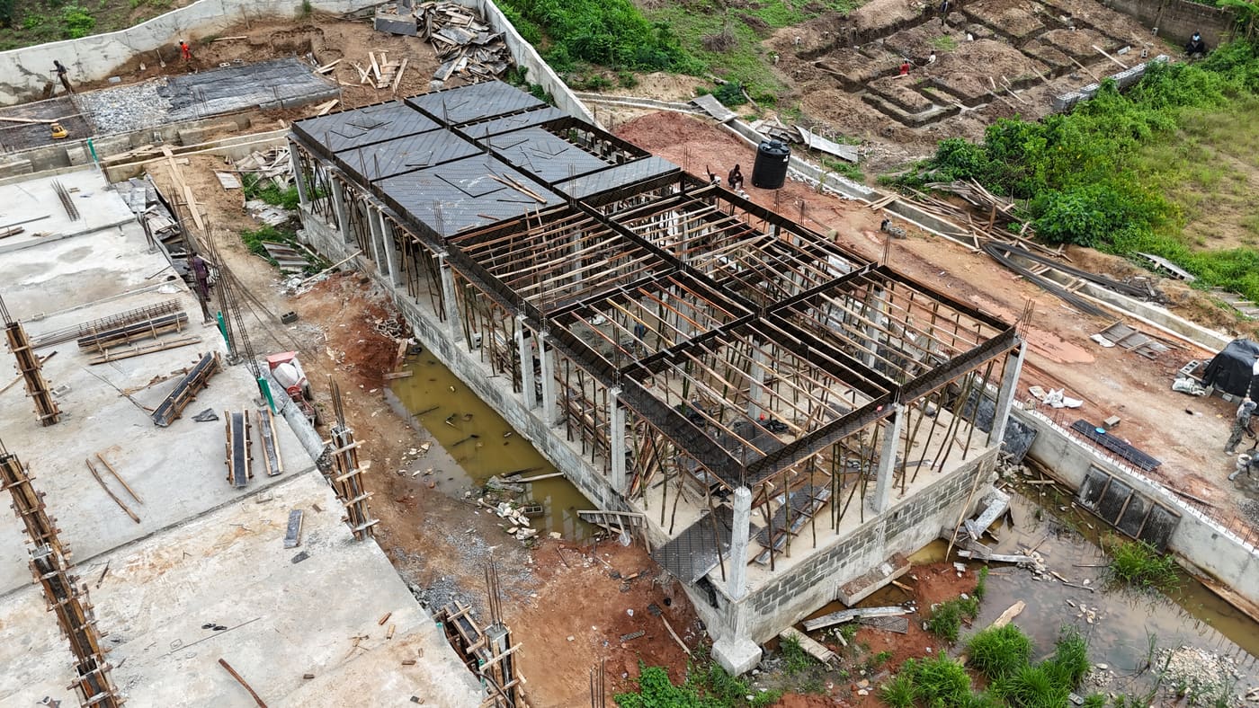 Ewang Estate under construction, 10 July 2025: the ground-floor slab cast and columns standing, with timber formwork and bamboo props carrying the first suspended slab as workers pour. Aerial view 4 of 4.