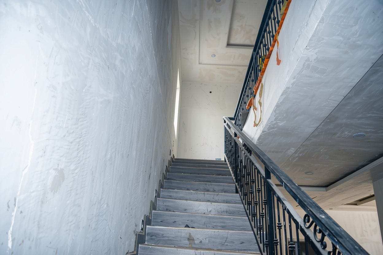 Staircase looking up through a finished home, with wood-effect tiled treads, a black scrolled balustrade and a coffered ceiling.
